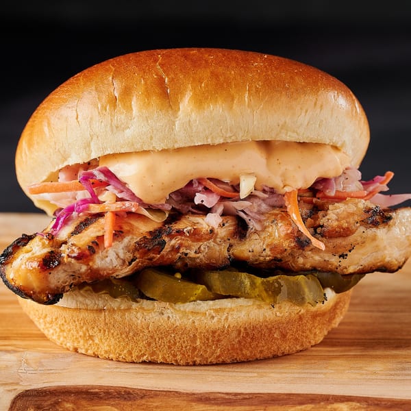 Grilled chicken sandwich with coleslaw, sauce, and pickles on a bun, on a wooden board.