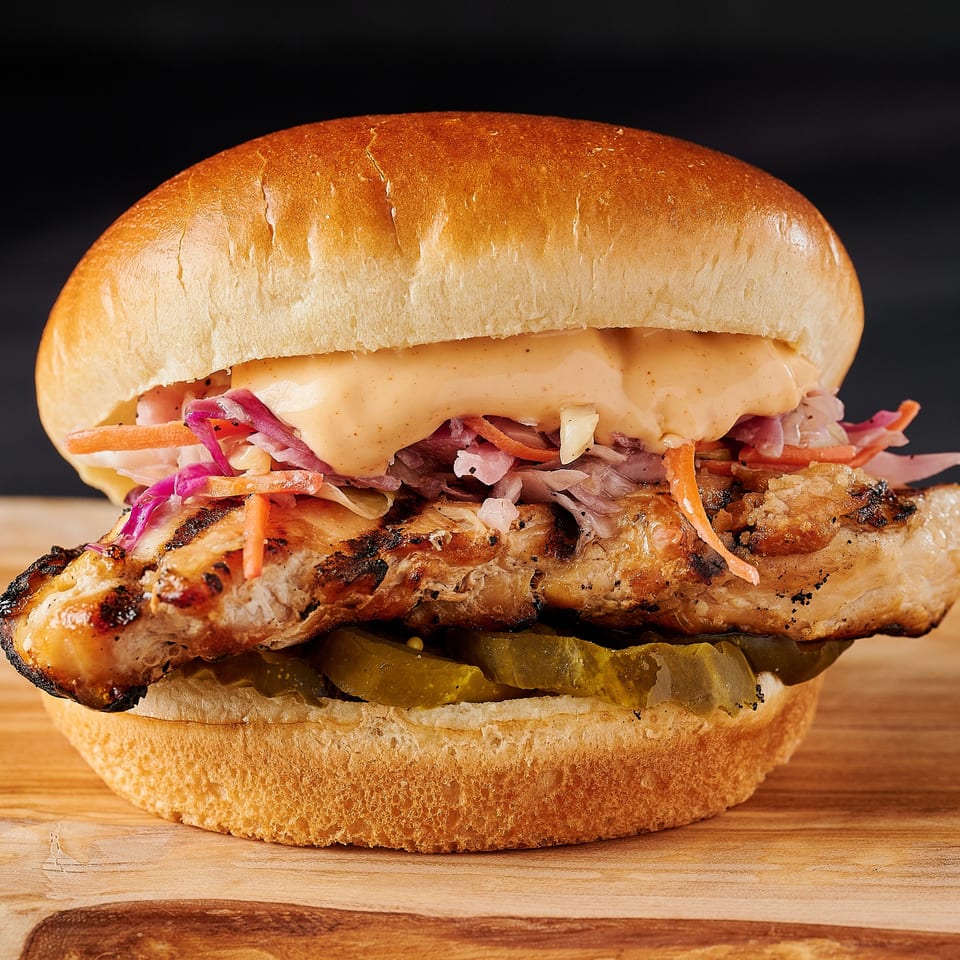 Grilled chicken sandwich with coleslaw, sauce, and pickles on a bun, on a wooden board.