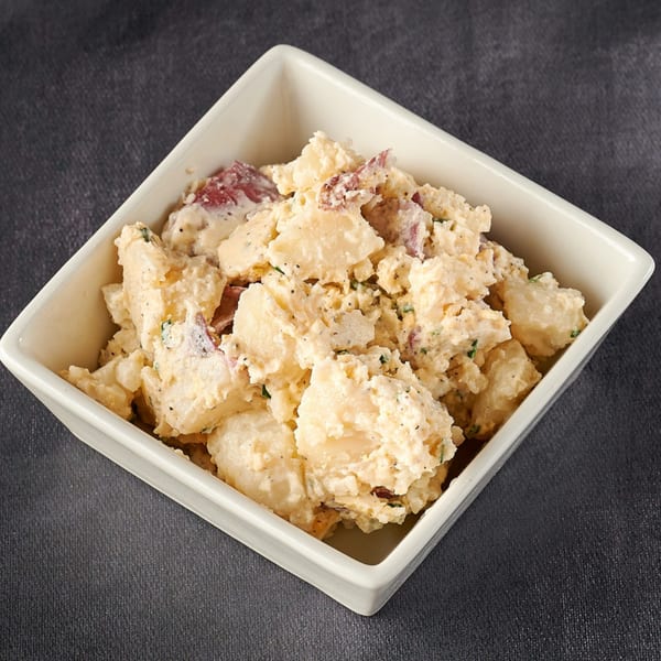 A square white bowl filled with creamy potato salad, featuring red potato skins and herbs.