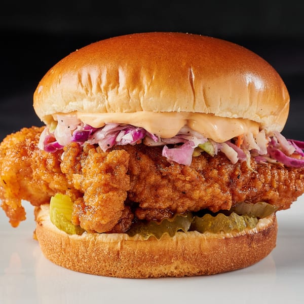 Hot Chicks House of Chicken's fried chicken sandwich with coleslaw, pickles, and sauce on a bun.